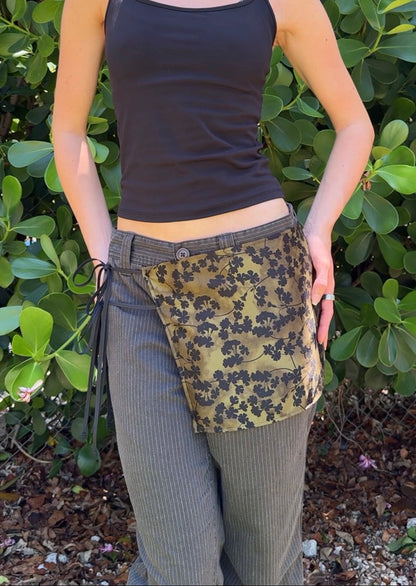 Person wearing a black tank top and green floral wrap skirt against a green bush background