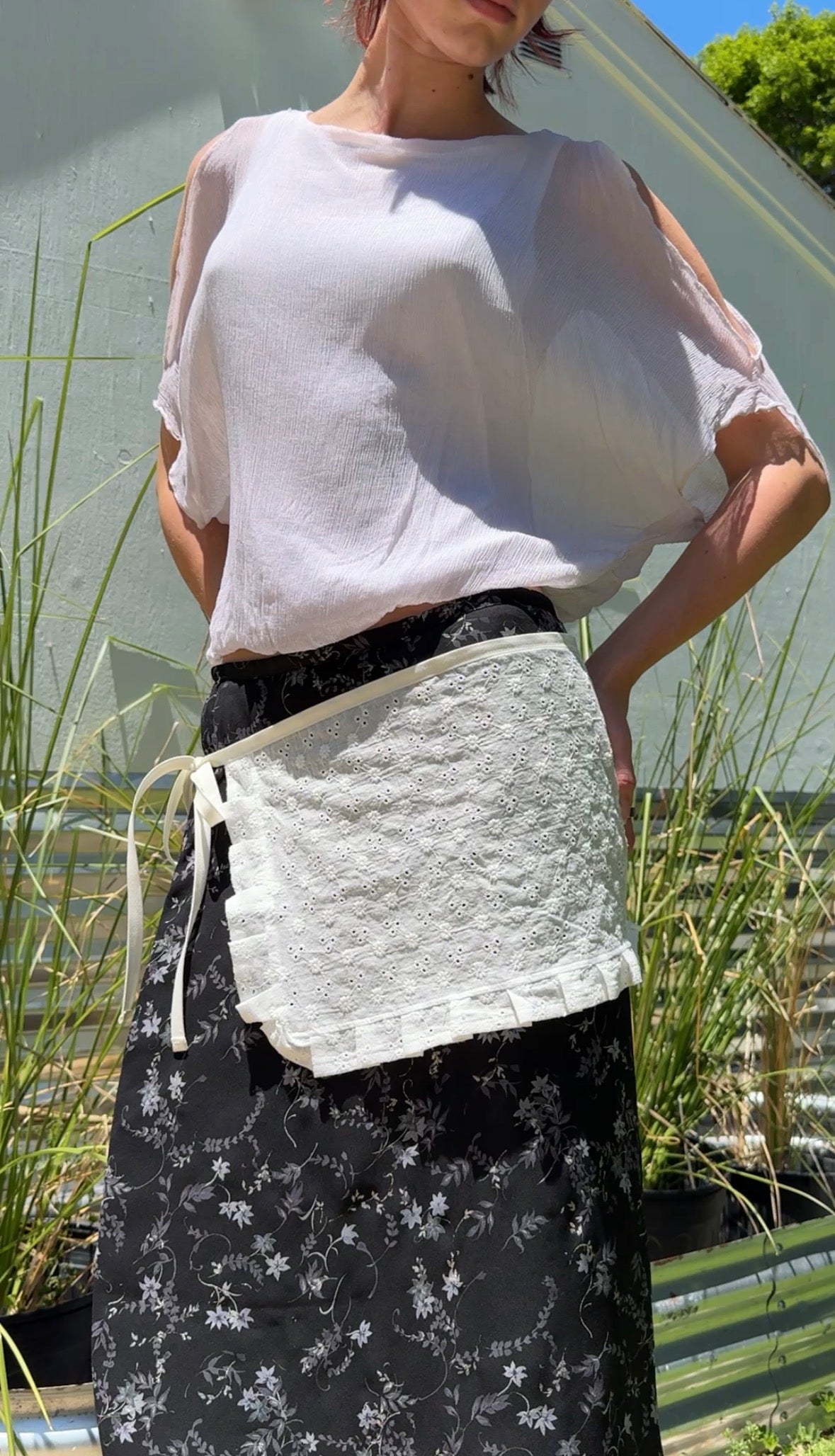 Person wearing a white apron over a light-colored shirt with a black floral skirt, standing outdoors.